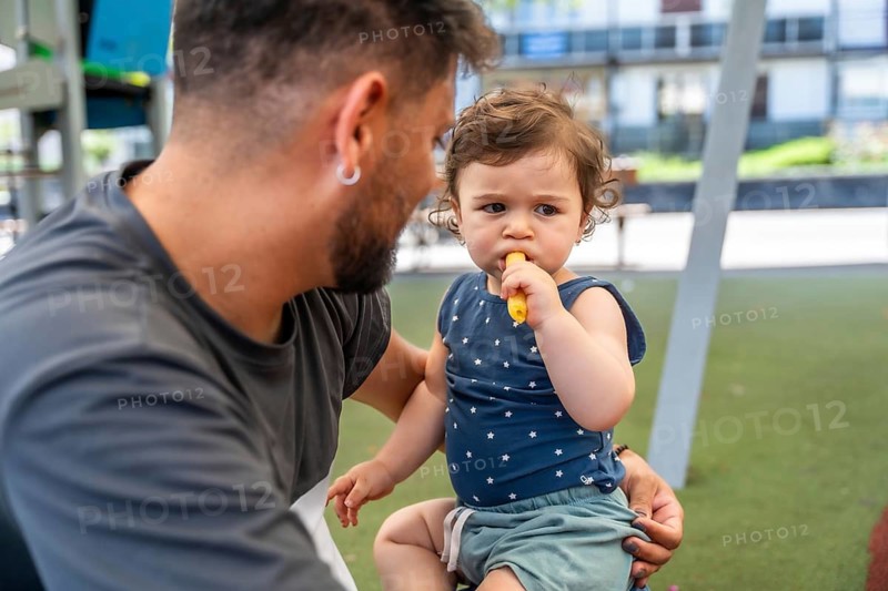 Father holding a young baby sitting on his lap, looking directly at the baby who is consuming a solid food item like a banana or carrot stick during a family outing in a public park