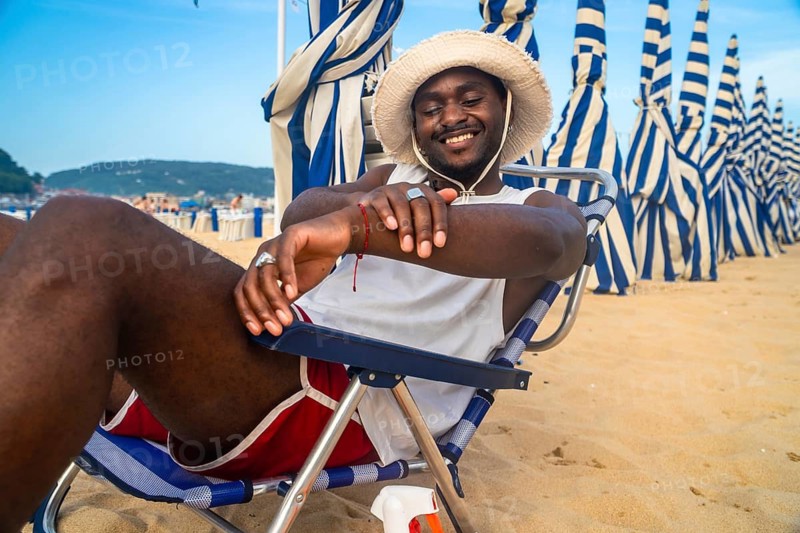 Smiling young man wearing a straw hat and sunglasses sitting comfortably in a striped beach chair on sandy beach, enjoying summer holiday with blue and white umbrellas in background