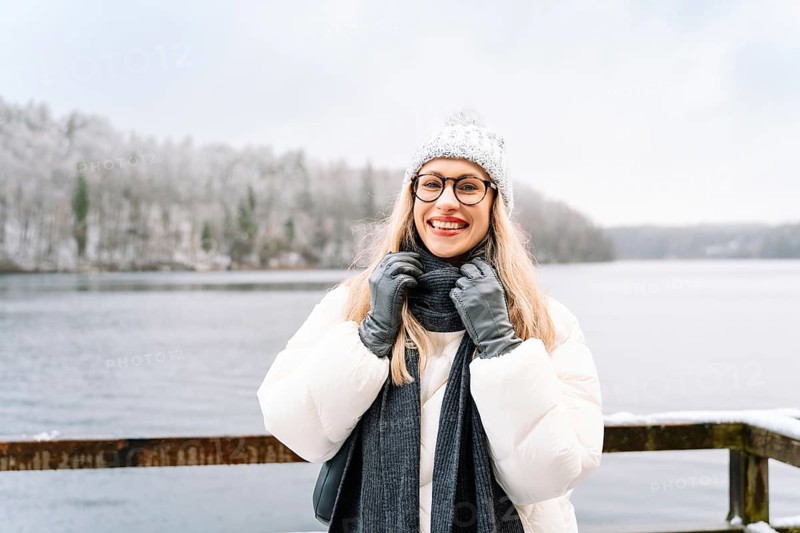 Smiling woman adjusting her scarf on a winter day near a snowy lake. A woman wearing a white winter coat, knitted hat, scarf, gloves and glasses stands outdoors near a snowy lake, adjusting her scarf with a cheerful, relaxed expression. Snowflakes rest on her clothing, creating a bright winter moment that highlights seasonal fashion, outdoor lifestyle and cold-weather travel