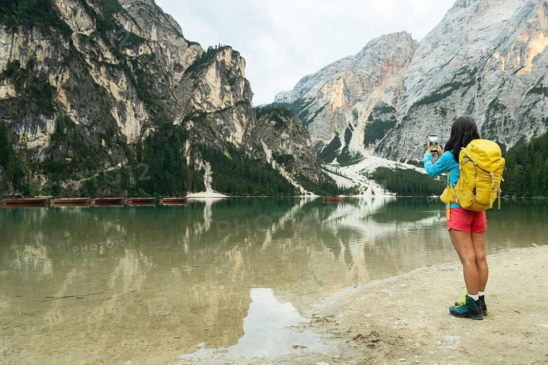 Woman hiker taking smartphone photo of Lago di Braies alpine lake turquoise water mountain reflections Dolomites Italy