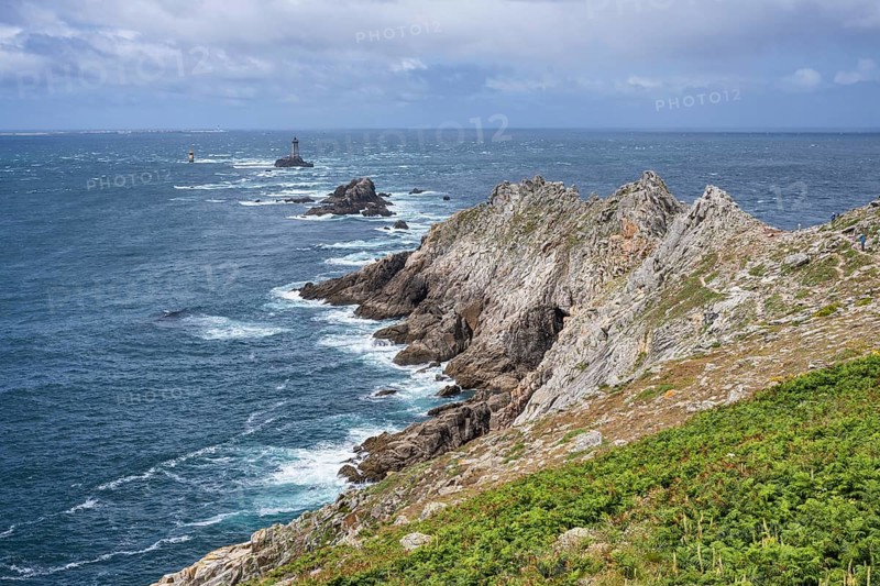 Steeply sloping, rocky cliffs of the Breton coast under blue skies with light clouds. The view extends far across the rugged landscape of rocks and barren vegetation to the deep blue Atlantic Ocean, with the Phare de la Vieille lighthouse on a rocky island in the background. Waves break foaming on the rugged coastline under bright daylight. Pointe du Raz, Finistère, Brittany, France