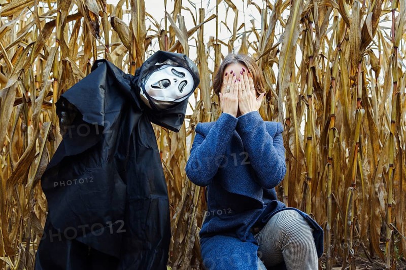 Woman covering her face with her hands near creepy scarecrow with mask of death in cornfield on Halloween