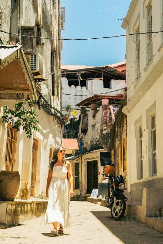 Woman tourist in white dress walks through beautiful Stone Town alley. Historic Zanzibar architecture and peaceful morning travel sightseeing.Popular travel destination Africa