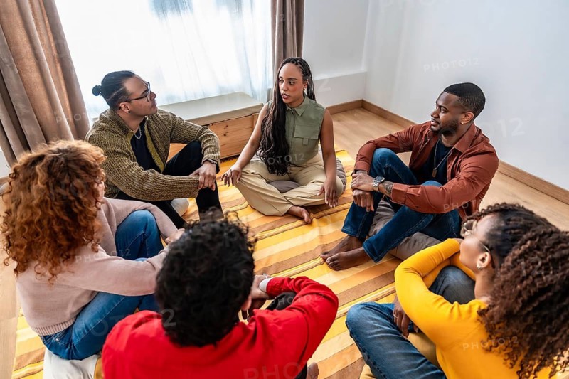 Diverse adults sitting in a circle on the floor, sharing experiences and actively listening, fostering community and mental well being in a supportive environment