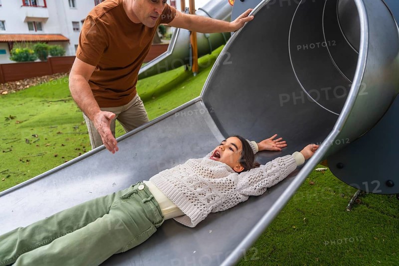 Expressive young girl with excited face sliding down a metal slide while father watches closely, sharing joyful outdoor playground bonding and carefree family fun at the park