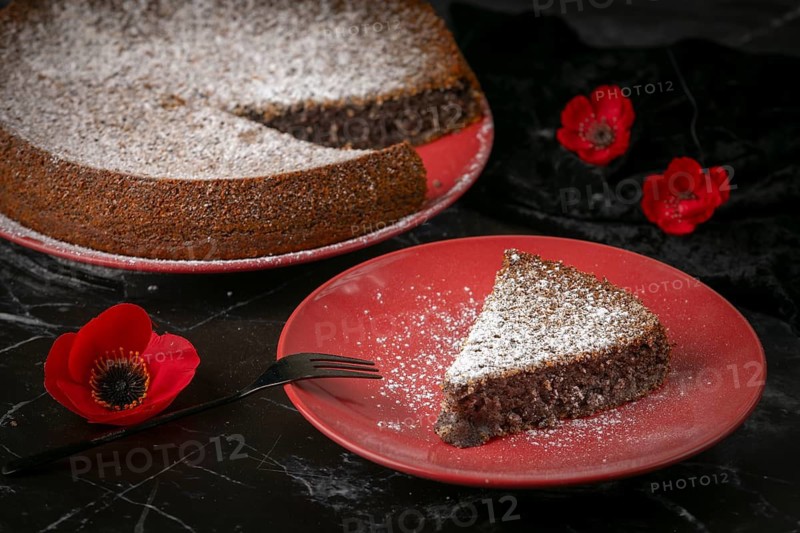 Sliced poppy seed cake on red plates with powdered sugar and poppies on a dark surface