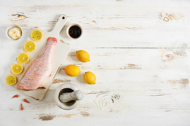 Raw red snapper is placed on cutting board next to sliced lemons and bowl of spices. On side are whole lemons and mortar with ingredients for seasoning. Cooking preparations take place here