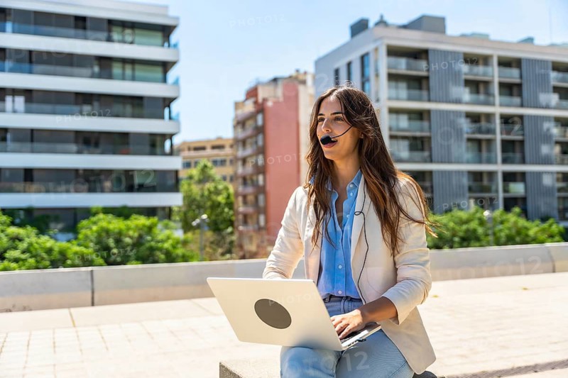 Woman having an online meeting or making a business call, working on her laptop with a headset, embracing the flexibility of remote work in an urban park or city setting during the day