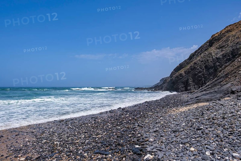 Pebble beach at the base of high cliffs, ocean waves rolling in, the sky is cloudless blue, Rogil, Faro, Portugal
