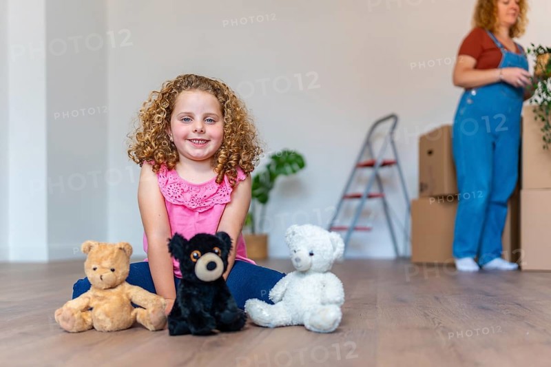 Young girl smiling and sitting on a wooden floor with teddy bears, experiencing the excitement of a new house while her mother unpacks belongings in the background