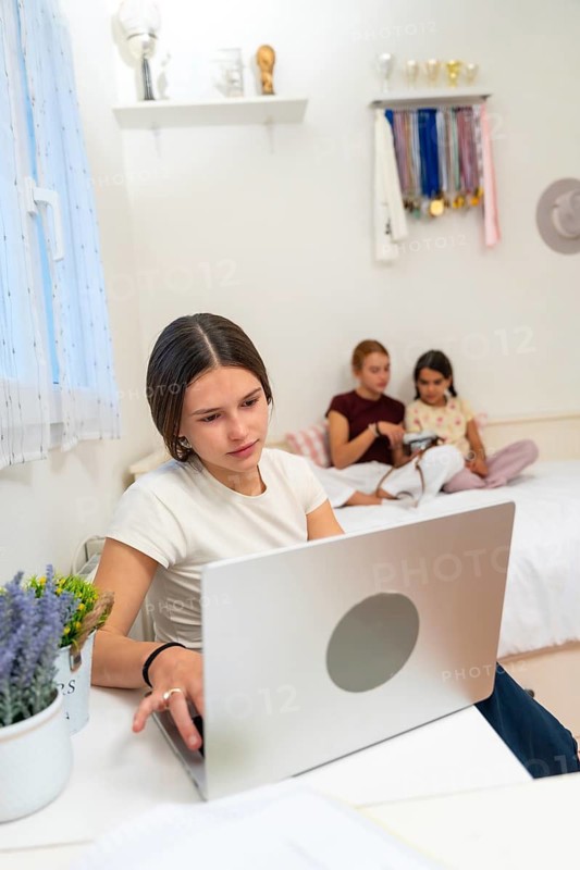 Teenager concentrating on her laptop at a desk, studying in her bedroom while her young sisters relax and play on the bed in the background, showing modern family life