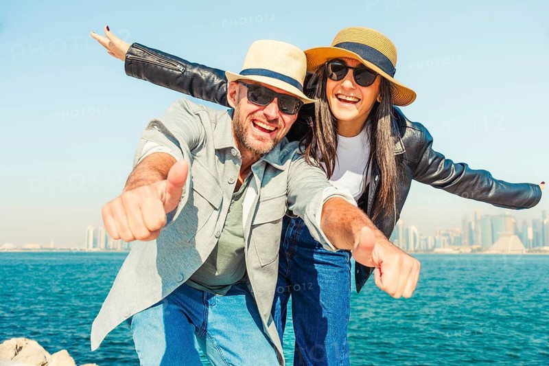 Happy tourist cute happy couple posing at bay. Man giving thumbs up, woman tongue out, sunglasses, hats, casual summer smart clothes. Skyline viewpoint by blue water on sunny day background