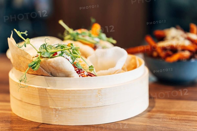 Freshly steamed bao buns in a traditional bamboo steamer, filled with vegetables, herbs, and sauce. Asian street food dish served in a modern restaurant setting