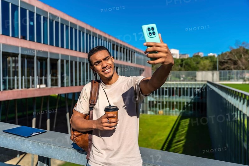 Young male student holding a coffee cup and smiling while taking a selfie on a sunny university campus, capturing a moment of contemporary college life and youthful technology usage