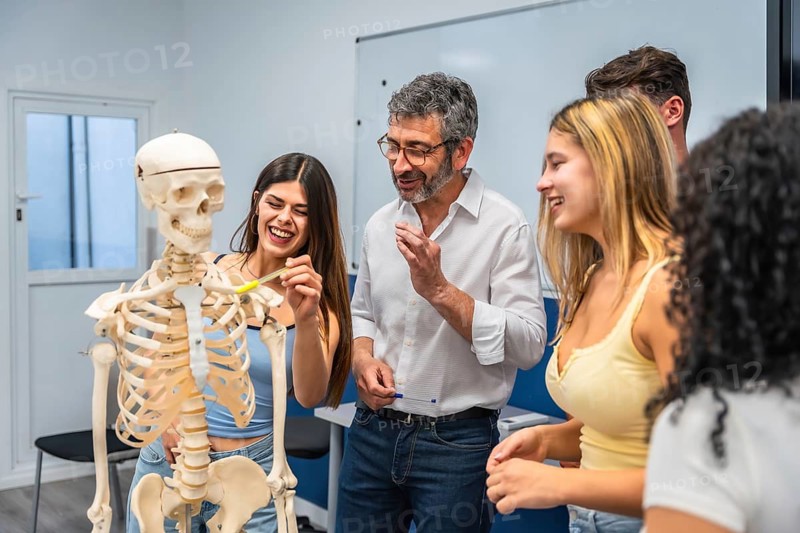 Young diverse students and an adult teacher engaging in a biology lesson, discussing the human skeleton model while pointing and smiling in a bright classroom setting