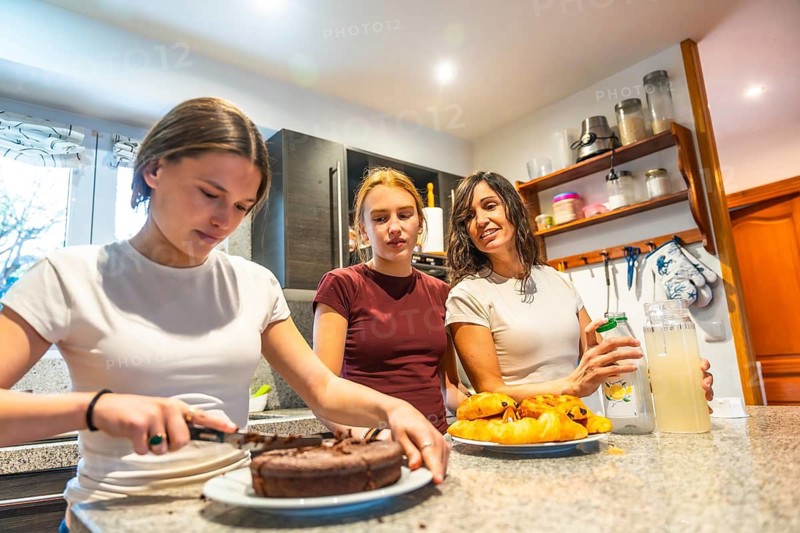 Three women of different generations sharing a happy moment in a bright kitchen, preparing a homemade chocolate cake and pouring fresh lemonade, symbolizing family bonding and togetherness