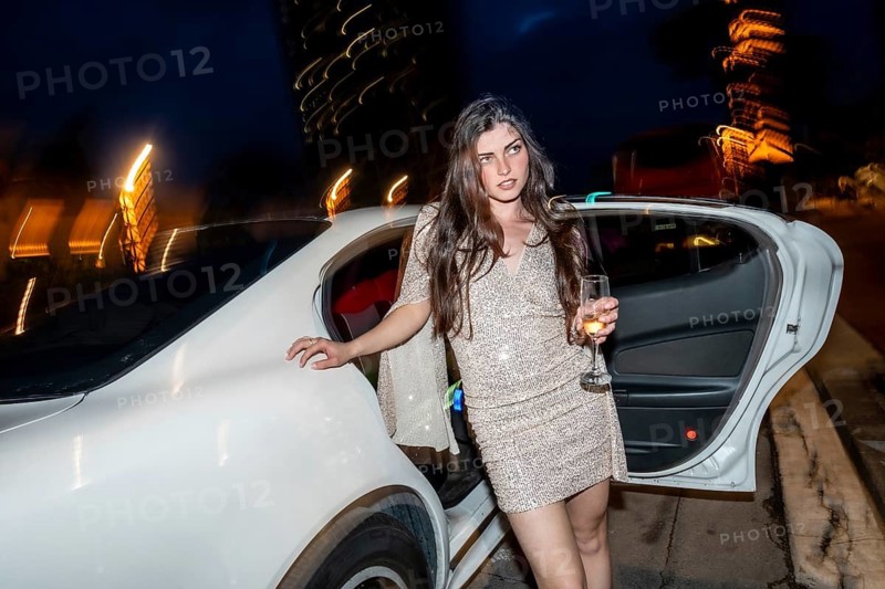 Young woman stepping out of a white limousine, holding a champagne glass and looking around during an elegant evening celebration, with blurred city lights in the background