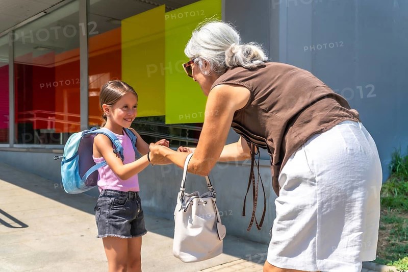 Grandmother is bending to greet her happy granddaughter. Wearing a backpack. Standing outside a modern school building after classes. Symbolizing family support. Education. And intergenerational bonds