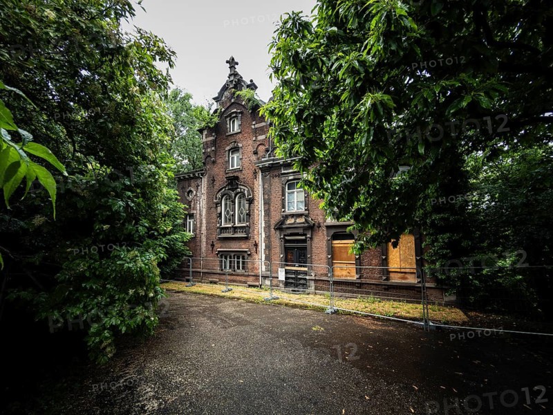 An old brick villa stands abandoned and boarded up with wooden panels behind a construction fence, surrounded by thick, green trees, Liège, Belgium