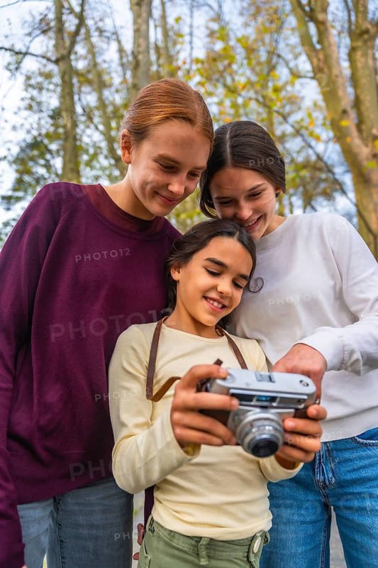 Three happy sisters looking at images on a vintage camera, sharing joy and bonding during a family outing in a park, learning and exploring the art of photography together in nature