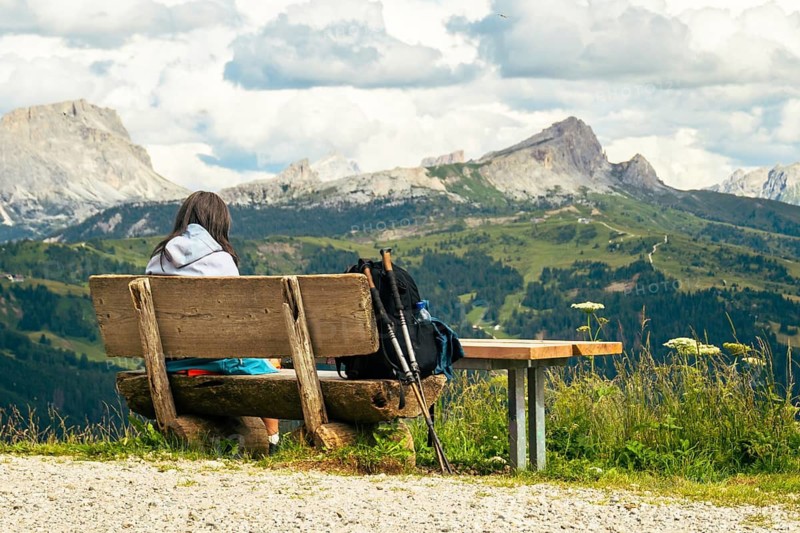 Caucasian woman sit alone on hand made wooden bench, admiring the majestic Dolomites mountain range in Italy. A peaceful moment of adventure, wellness, and scenic trekking in South Tyrol
