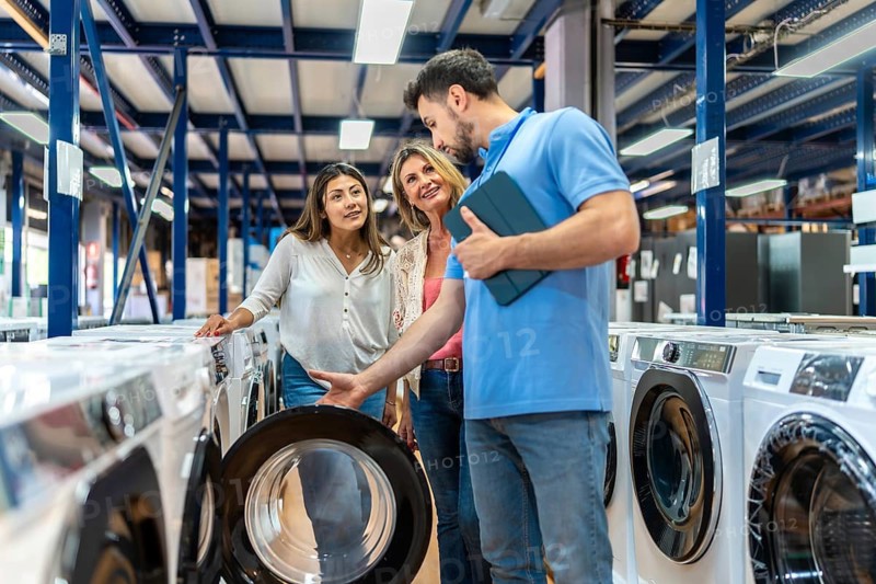 Sales associate helping a mother and daughter select a new washing machine at an appliance store, providing information and guidance on different models and features for their home