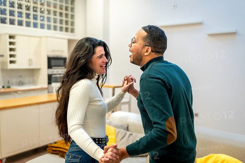 Happy diverse couple holding hands and dancing intimately in a modern open plan living room and kitchen, enjoying a joyful moment of connection and movement at home
