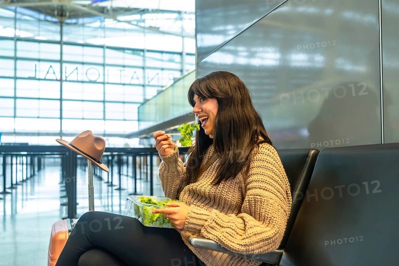 Young female traveler with dark hair sitting on a black bench in a modern airport terminal, enjoying a fresh green salad from a plastic container before departure, luggage nearby