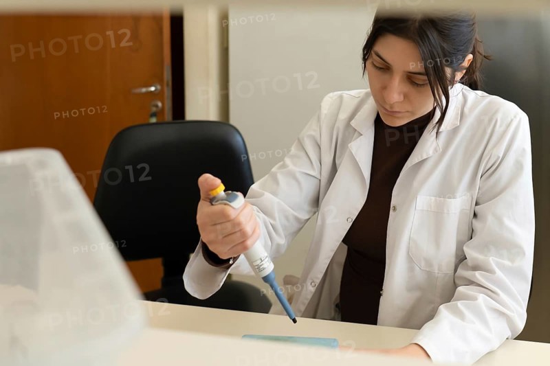 Young female scientist in white lab coat carefully using a micropipette at a desk in a bright laboratory, with a blurred foreground and office chair in the background