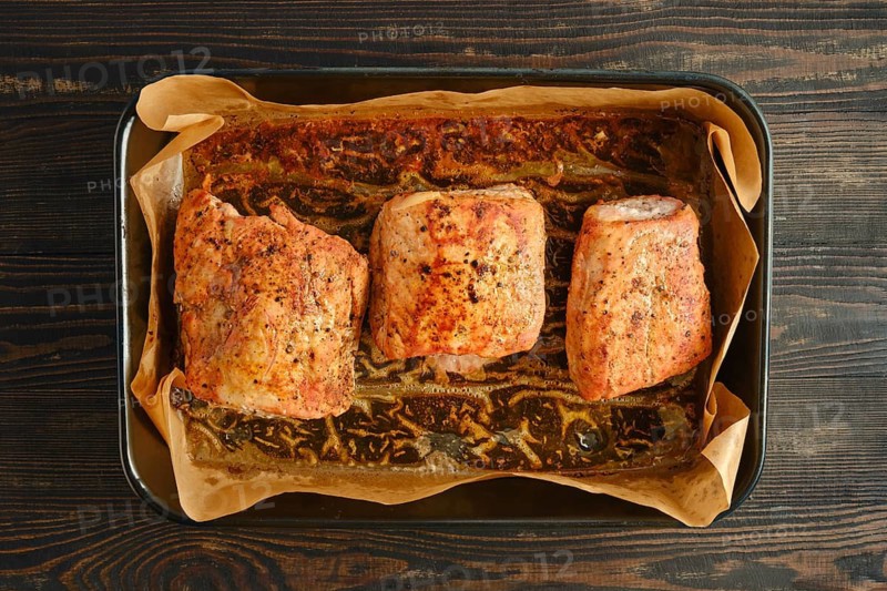 Pork fillets on baking sheet with parchment paper. Seasoning and herbs are visible on meat. Countertop is made of dark wood. Cooking is taking place in kitchen during afternoon