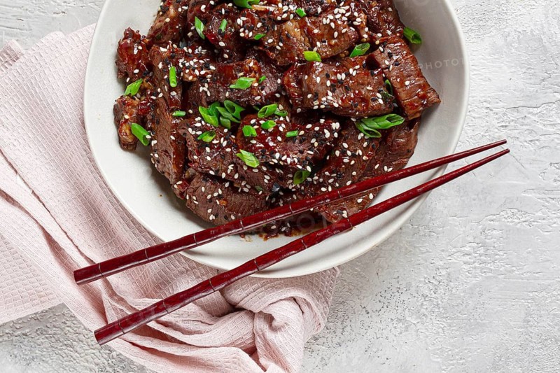 Crispy beef, with green onions and sesame seeds, in a bowl with chopsticks, on a textured surface, on a light background, no people