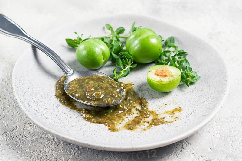 A spoon with tkemali sauce, on a plate, with cherry plum and mint, top view, no people