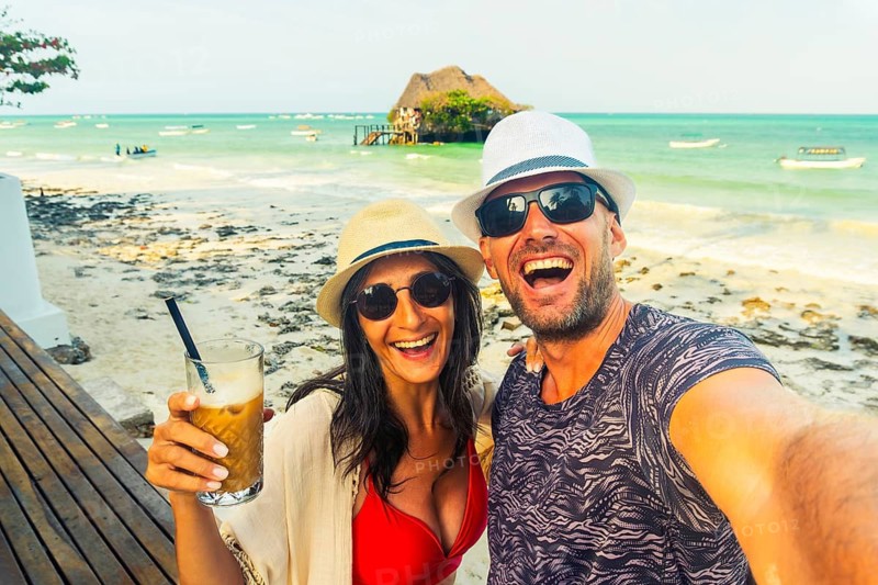 Happy couple smiles at camera takes playful selfie on a tropical beach with famous attraction rock restaurant behind. Authentic joy, laughter, cheers holiday maker and summer vacation vibes in paradise