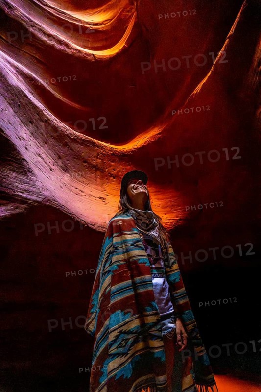 Woman standing inside antelope canyon wearing a colorful poncho, looking up at the sandstone walls illuminated by soft light, enjoying the natural beauty