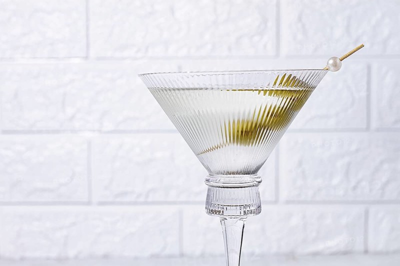 A clear glass cocktail sits on a white marble surface. Three green olives are placed on a skewer resting inside the glass. The light shines on the glass, highlighting its shape