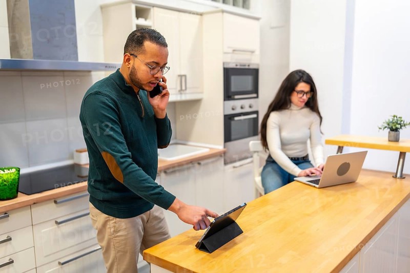 Young man talking on phone and using tablet while standing in the kitchen as a woman works on her laptop at the counter, illustrating a modern remote work lifestyle