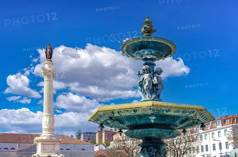 An ornate bronze baroque fountain near the monumental Column of Pedro IV under a vibrant blue sky with clouds at the historic Rossio Square in Lisbon Portuga