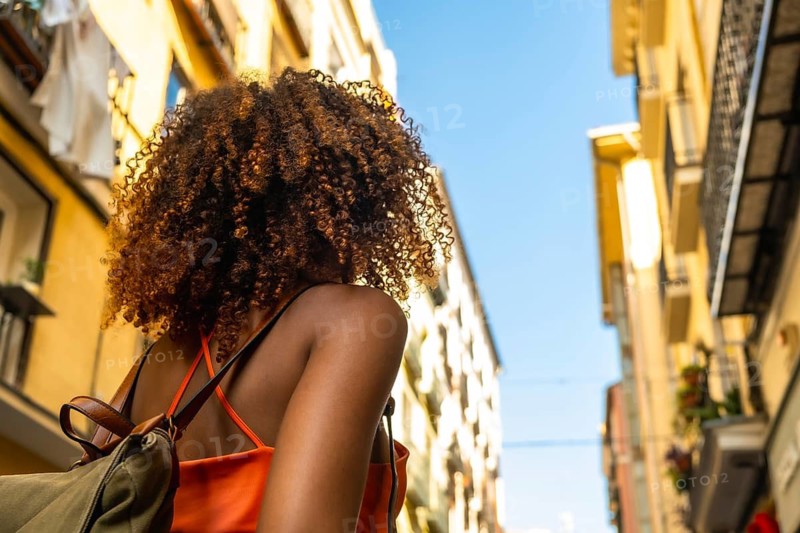 Woman with curly hair wearing an orange top and backpack walking through a narrow historic city street, exploring urban architecture under a clear blue sky