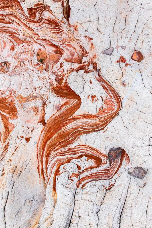 A close-up aerial photograph capturing the intricate patterns and textures of abstract red and white rocks. The swirling lines and cracked surface create a unique natural artwork