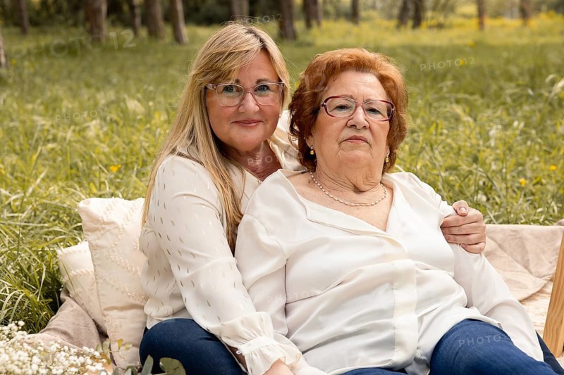 A mother and daughter share a loving moment in a serene outdoor setting. Sitting together on a soft blanket, their warmth and connection convey deep affection and lifelong companionship