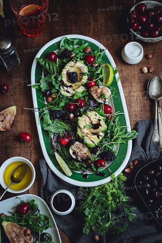 A vibrant salad featuring avocado, figs, cherries, and mixed greens on a rustic wooden table. The fresh ingredients create a colorful and appetizing presentation