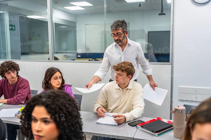 Education professional handing out exam papers to a diverse group of focused young adult students in a modern classroom, illustrating assessment, instruction, and academic learning preparation