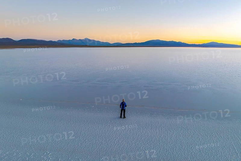 A lone figure stands on the expansive salt flats of Salinas Grandes in Salta, Argentina, as the sun sets, casting a gentle glow over the tranquil landscape and distant mountains