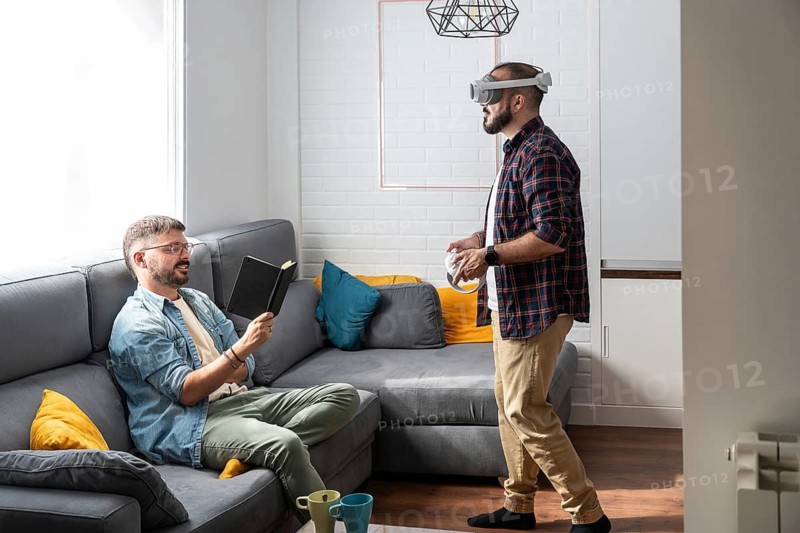 A gay couple enjoys a relaxing day at home, one reading on the couch while the other explores virtual reality. Their cozy living room is a mix of modern and comfortable elements