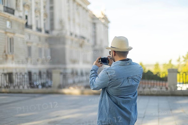 A Latino transgender man in a denim jacket and hat captures photographs of city architecture The image conveys a sense of exploration and pride in identity