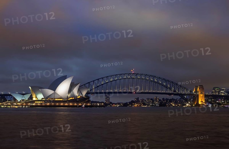 Captivating night view of Sydney Harbor, featuring the illuminated Opera House and Harbour Bridge under a cloudy sky and lights reflecting on the still waters below