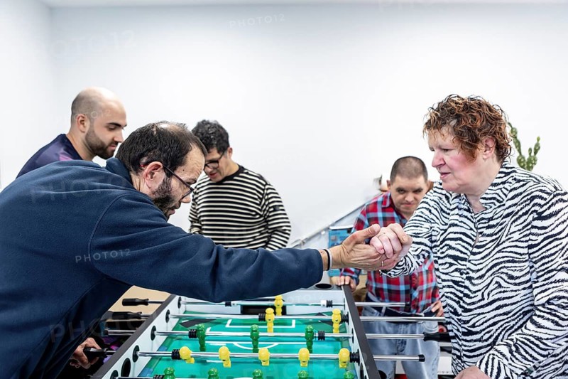 Residents of a mental health facility enthusiastically engage in a foosball game, showcasing camaraderie and therapeutic recreation. The scene emphasizes community and well-being