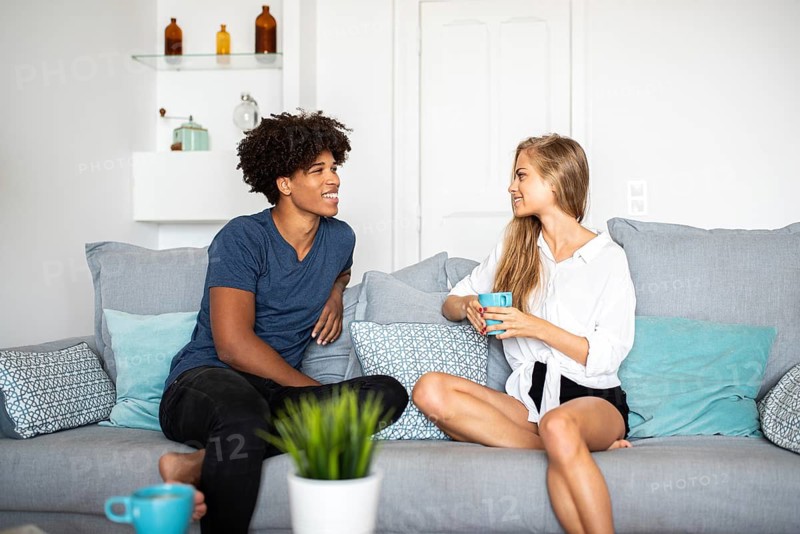Two friends sit in a modern living room, enjoying coffee and chatting. The cozy setting features a light grey sofa, decorative pillows, and a plant, creating a warm atmosphere