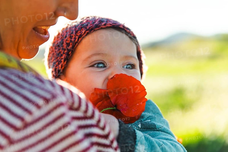 A mother holds her baby who is playfully biting a vibrant red flower. The image captures a sunny day outdoors in Samalus, Barcelona, Spain, evoking warmth and joy