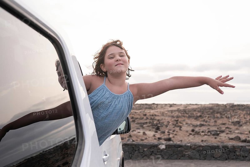 A young girl joyfully leans out of a car window, feeling the breeze as she embraces the spirit of a summer road trip. The rocky landscape adds a sense of adventure